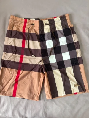 Rutiga badbyxor - Snygga badbyxor med klassiskt rutigt mönster i beige, brunt, svart, vitt och röda detaljer. De har elastisk midja med dragsko och en bakficka med knapp. Perfekta för stranden eller poolen, tillverkade i snabbtorkande material.