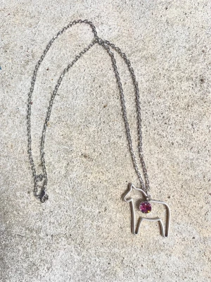 Silverfärgat halsband med dalahäst - Unikt halsband i silverfärgad metall med ett hänge format som en dalahäst. På hästen sitter en rund, rosa pärla som ger en snygg kontrast. Kedjan är tunn och smidig, perfekt för dig som gillar stilrena accessoarer med en twist. Den rosa stenen är oktober månadssten. Kedjan är 60 cm lång