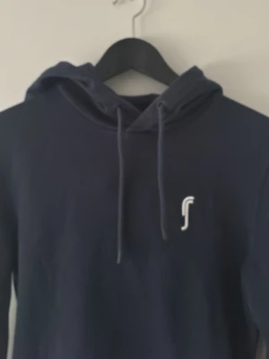 Mörkblå hoodie med vit logga - Snygg mörkblå hoodie med huva och dragsnören. Framtill finns en liten vit logga på bröstet och en stor magficka. Hoodien har långa ärmar och är tillverkad i mjukt material, perfekt för en avslappnad stil.