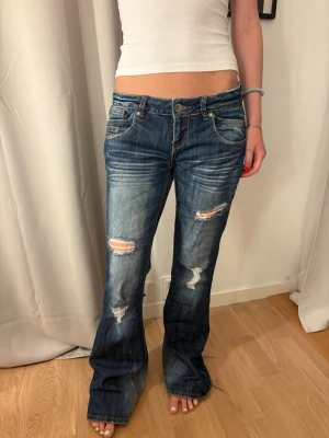 Blå slitna bootcut jeans med nitar - Säljer ett par blå bootcut jeans med slitningar och hål på knäna. Jeansen har dekorativa nitar och mönster på bakfickorna samt ljusa tvättade partier. Klassisk femficksmodell med låg midja och dragkedja. Perfekta för en trendig och avslappnad look.