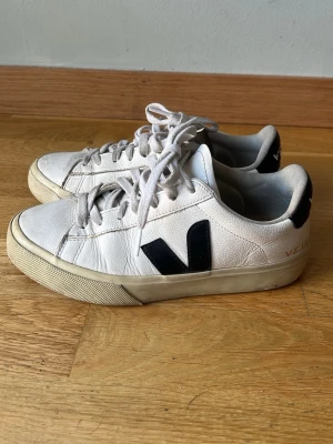 Vita Veja sneakers med svart V-logga - Snygga vita sneakers från Veja med svart V-logga på sidorna. Skorna har klassisk låg modell, vita skosnören och beige sula. Tillverkade i läder med textildetaljer på insidan. Perfekta för en stilren och trendig look.