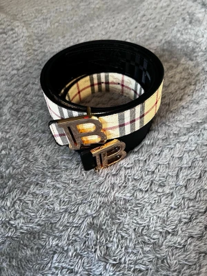 Rutig Burberry skärp med BB-spänne - Snyggt rutig Burberry skärp i klassiska färger med ett guldigt BB-spänne framtill. Skärpet har beige, svarta och röda ränder och är gjort i skinn. Perfekt accessoar för att lyfta din outfit.