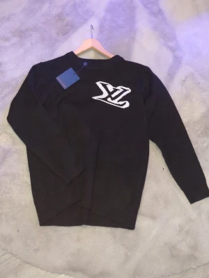 Svart stickad tröja med LV-logga - Snygg svart tröja från Louis Vuitton med stor vit LV-logga på bröstet. Tröjan har rund halsringning och långa ärmar. Perfekt för dig som vill ha en stilren och exklusiv look.