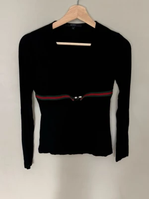 Gucci Svart långärmad topp med randig detalj - Svart långärmad topp med smal passform och en randig detalj i rött och grönt runt midjan. Toppen har en enkel design med rund halsringning och är perfekt för dig som gillar stilrena plagg med en twist.0765