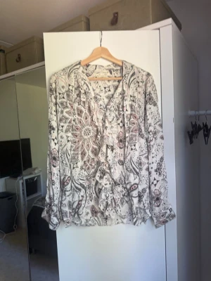 Mönstrad skjorta i vitt och brunt - Säljer en vacker mönstrad skjorta i vitt och brunt med inslag av svart. Blusen har långa ärmar, knappar framtill och ett bohemiskt mönster med blommor och paisley. Perfekt till både vardag och fest! Från märket Odd Molly i storlek 3 som enligt deras storlekguide motsvarar en storlek L. Mycket fint skick, jag hittar inga synliga anmärkningar eller defekter. 