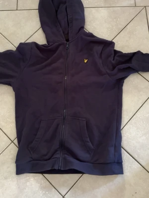 Mörkblå hoodie från Lyle & Scott - Säljer en mörkblå hoodie från Lyle & Scott med dragkedja framtill och två stora fickor. Klassisk gul logga på bröstet och skön huva. Perfekt för dig som gillar enkel och stilren streetwear.