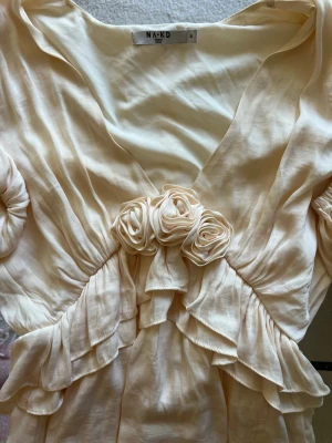 Beige volangblus  - Romantisk beige blus från NA-KD med djup v-ringning, volangdetaljer och dekorativa tygrosor framtill. Blusen har långa, luftiga ärmar och ett mjukt, glansigt material som ger en lyxig känsla. Storlek 34💞