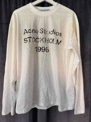 Acne Studios  - Säljer en stilren, långärmad t-shirt från svenska Acne Studios med det ikoniska ”Stockholm 1996”-trycket på bröstet. Tröjan har en avslappnad passform, rund hals och är tillverkad i en mjuk, högkvalitativ bomullsmix som faller väldigt snyggt. En perfekt klassiker till basgarderoben. STORLEK M 