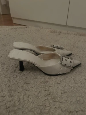 Vita spetsiga kitten heels med stor spänne - Säljer ett par vita kitten heels med spetsig tå och öppen häl. Skorna har en smal klack och en stor dekorativ spänne i silver framtill. Materialet ser ut som konstläder med en lätt glansig finish. Perfekta för dig som vill sticka ut med en trendig och elegant look.