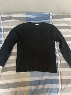 Svart stickad tröja med rund hals - En enkel och stilren svart stickad tröja med rund hals och långa ärmar. Tröjan har ribbade muddar vid ärmslut och nederkant, vilket ger en snygg och klassisk look. Perfekt att matcha med jeans.