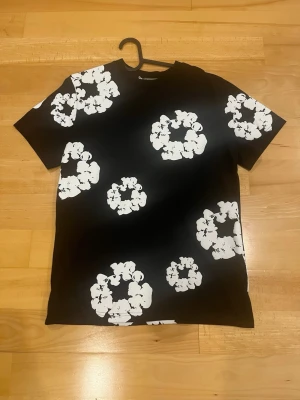 denim teats t shirt - Cool svart t-shirt med stora vita blommor i ett grafiskt mönster över hela plagget. T-shirten har rund hals och korta ärmar. Perfekt för dig som vill sticka ut med en unik och trendig look. Tillverkad i mjukt material som känns skönt mot huden.