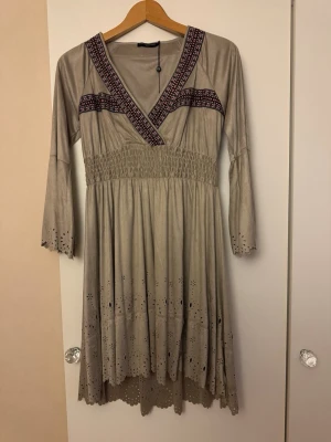  Klänning med broderi och v-ringning - Klänningen har trekvartsärm med utsvängda ärmslut och fina hålbroderier längs ärmar och fåll. Smock i midjan för skön passform och mjukt, tunt material. Bakstycket är lite längre än framstycket. Klänningen är enbart provad.