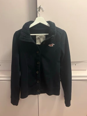 Mörkblå kofta från Hollister - Mysig mörkblå kofta från Hollister med ribbad krage och knappstängning framtill. Klassisk logga broderad på bröstet och långa ärmar. Perfekt för lager på lager och enkel att matcha med olika outfits.