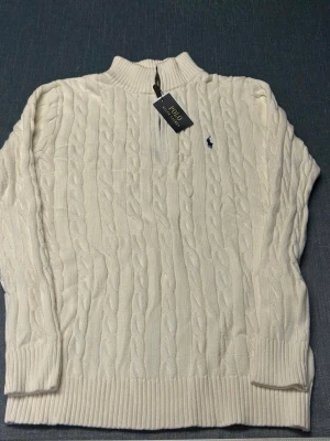 Vit kabelstickad tröja Polo Ralph Lauren - Kabelstickad vit tröja från Polo Ralph Lauren med hög krage och ribbade muddar. Klassisk design med broderad logga på bröstet. Perfekt för dig som gillar stilrena och tidlösa plagg i mjukt material.