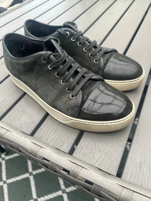 Lanvin skor | 43 - Snygga svarta sneakers med mockaliknande material på sidorna och glansig lackad tå. Skorna har svarta snören och vit sula som ger en clean kontrast. Perfekta för dig som gillar stilrena och moderna skor. Har haft en sula i som gör att det ser lite användt ut i sulan