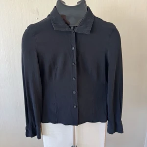 Versace Svart klassisk blus med krage - Stilren svart blus med klassisk krage och knäppning framtill. Blusen har långa ärmar och är tillverkad i ett tunt, lätt material som ger en snygg siluett. Perfekt att matcha med både jeans och kjol för en trendig look.0336