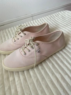 Ljusrosa tygsneakers med vita snören, Gant - Säljer ett par ljusrosa sneakers i tyg med vita snören och vita sulor. Skorna har en enkel och klassisk design med metallöljetter och en liten broderad logga på sidan. Perfekta för en avslappnad och stilren look.
