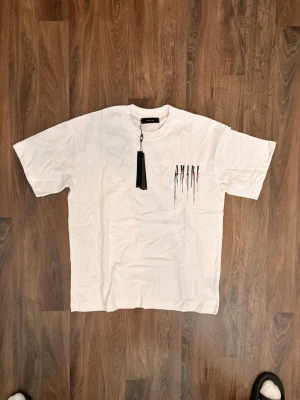 Vit AMIRI t-shirt med färgstänk - Säljer en vit t-shirt från AMIRI med coolt tryck framtill där loggan är dekorerad med färgstänk i svart, blått och orange. T-shirten har klassisk passform och korta ärmar. Perfekt för dig som vill sticka ut med en unik streetwear-look.