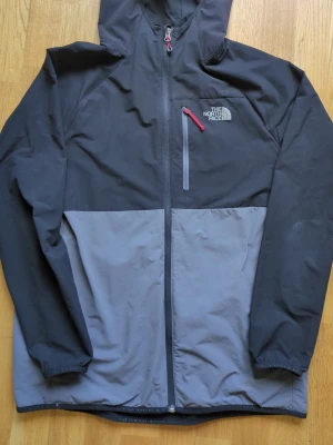Svart och grå vindjacka The North Face - Snygg vindjacka från The North Face i svart och grått med huva och hel dragkedja. Jackan har en bröstficka med röd dragkedja, elastiska muddar och logga på bröstet och ryggen. Perfekt för blåsiga dagar och outdoor-äventyr. Köptes på JD Sports för 899kr. Säljes för 400kr. Pris kan diskuteras vid snabb affär. Tvättas innan köp.