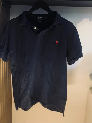Marinblå piké från Polo Ralph Lauren - Klassisk marinblå pikétröja från Polo Ralph Lauren  och den ikoniska röda logon broderad på bröstet. Tillverkad i mjuk bomull och har krage med knappslå. Perfekt till en avslappnad men stilren look. Storlek L men passar M, pris kan diskuteras vid snabb affär 
