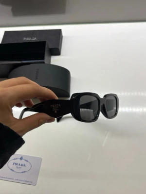 Svarta solglasögon från Prada - Snygga svarta solglasögon från Prada med rektangulära bågar och mörka glas. Klassisk och stilren design med diskret Prada-logga på sidan. Perfekta för dig som vill ha en trendig accessoar med exklusiv känsla.