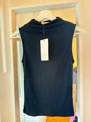 Svart ärmlös polotopp från Zara - Snygg svart polotopp från Zara med ärmlös design och hög hals. Toppen är enkel och stilren, perfekt att matcha med olika outfits. Tillverkad i mjukt material som sitter skönt mot kroppen.
