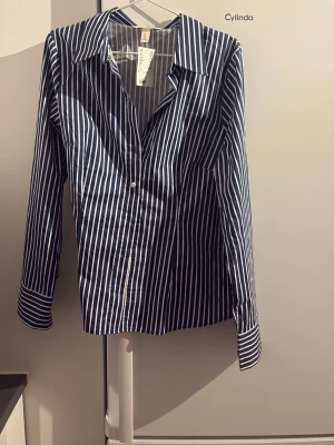 Blåvit randig skjorta med knappar - Snygg blå skjorta med vita vertikala ränder och klassisk krage. Skjortan har långa ärmar och knäppning framtill. Perfekt till jeans eller kjol för en clean look. Figursydd.