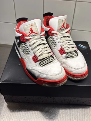Air Jordan 4 Retro Fire Red  - Säljer ett par Air Jordan 4 Retro Fire Red sneakers med vit ovandel i läder, röda och svarta detaljer samt meshpaneler på sidorna. Skorna har klassisk Jumpman-logga på plösen och Nike Air-logga på hälen. Röd innersula och svart-vit-röd yttersula.