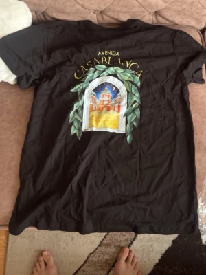 Svart t-shirt med Casablanca-tryck - Säljer en svart t-shirt med färgglatt tryck av en byggnad och texten 'Avenida Casablanca' på både fram- och baksidan. T-shirten har korta ärmar och är i mjukt bomullsmaterial. Perfekt för dig som gillar coola grafiska motiv.