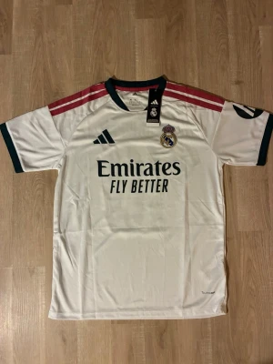  Real Madrid Hemmatröja 26/27 Mbappe - Snygg vit Real Madrid Hemmatröja med svarta och rosa detaljer. Tröjan har klubbmärke på bröstet och sponsortryck framtill. Korta ärmar med gröna muddar och rosa ränder på axlarna. Perfekt för fotbollsträning eller match. HELT NY!