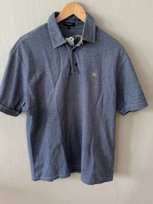 Blå pikétröja från Burberry - Klassisk blå pikétröja från Burberry med korta ärmar och krage. Tröjan har två knappar framtill och en liten diskret logga på bröstet. Perfekt till jeans eller shorts för en clean look.0088