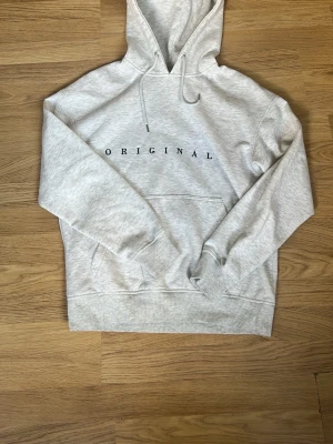 Ljusgrå hoodie från Jack & Jones - Snygg ljusgrå hoodie från Jack & Jones med trycket 'ORIGINAL' över bröstet. Klassisk känguruficka framtill, dragsko i huvan och mjukt material. Perfekt för en avslappnad och trendig stil. M/38/10