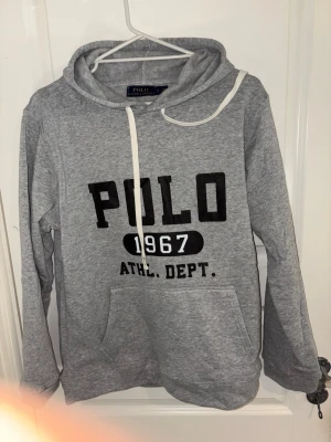 Grå hoodie från Polo Ralph Lauren - Säljer en grå hoodie från Polo Ralph Lauren med svart tryck 'POLO 1967 ATHL. DEPT.' på bröstet. Hoodien har huva med vita snören och en stor magficka. Tillverkad i mjukt material, perfekt för chill dagar.