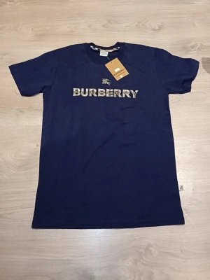 Mörkblå Burberry t-shirt - Snygg mörkblå t-shirt från Burberry med det klassiska Burberry-logotypen broderad i vitt och rutigt mönster på bröstet. T-shirten har rund hals och korta ärmar. Perfekt för dig som vill ha en stilren och exklusiv look.