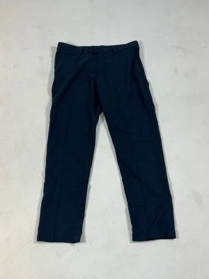 Mörkblå chinos från J.Lindeberg - Snygga mörkblå chinos från J.Lindeberg med klassisk rak passform och diskreta sidofickor. Tillverkade i ett mjukt och slitstarkt material som ger en stilren look. Perfekta för dig som vill ha en clean och modern stil eller ett par feta golf byxor storlek 32/32