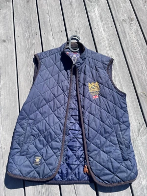 Marinblå quiltad dunväst Morris - Snygg marinblå quiltad dunväst från Morris med guldfärgad brodyr och brittisk flagga på bröstet. Västen har ståkrage, två fickor fram och bruna kantband. Perfekt för lager-på-lager och ger en klassisk preppy stil.