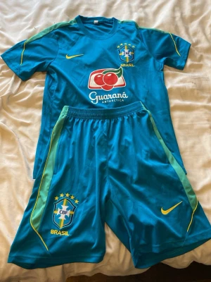 Blått Brasilien fotbollsställ Nike - Säljer ett blått Brasilien fotbollsställ från Nike med matchande tröja och shorts. Tröjan har CBF-logga, sponsortryck och gula detaljer. Shortsen har elastisk midja, CBF-märke och gula Nike-loggor. Perfekt för träning eller match!