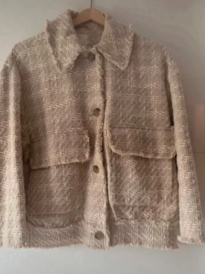 Beige tweed cardiganjacka med fickor - Säljer en beige cardiganjacka i tweed med grov struktur och fiskbensmönster. Jackan har stora framfickor, knappar framtill och en klassisk krage. Perfekt för dig som gillar en trendig och avslappnad look. Använd 2 gånger, kan passa även M.