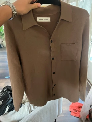 Brun kofta/ overshirt från Samsøe Samsøe - Snygg brun kofta/ overshirt från Samsøe Samsøe med klassisk krage, svarta knappar och en bröstficka. Tröjan har lång ärm och är tillverkad i ett mjukt, stickat material som ger en avslappnad men stilren look.
