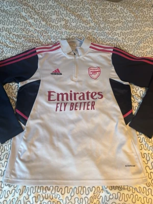 Arsenal träningströja Adidas långärmad - Snygg långärmad träningströja från Arsenal i vitt och marinblått med rosa detaljer. Tröjan har halv dragkedja, klubbmärke och Adidas-logga på bröstet samt 'Emirates Fly Better' tryckt framtill. Tillverkad i funktionsmaterial Aeroready. Det är storlek S men skulle säga att den är stor i storleken eftersom att de är en fotbolls tröja!