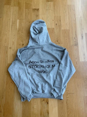 Grå hoodie från Acne Studios - Snygg grå hoodie från Acne Studios med tryck på ryggen där det står 'Acne Studios STOCKHOLM 1996'. Klassisk passform med huva och ribbade muddar vid ärmslut och nederkant. Perfekt för en avslappnad och trendig look.