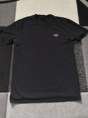 Svart t-shirt från Prada - Säljer en svart t-shirt från Prada med klassisk passform och rund hals. På bröstet sitter en liten trekantig Prada-logga i rött och vitt. Perfekt för dig som gillar stilrena och exklusiva plagg.