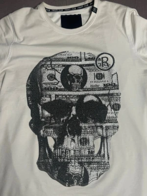 Vit t-shirt med döskalle från Philipp Plein - Säljer en vit t-shirt från Philipp Plein med ett stort svart tryck av en döskalle och sedlar på framsidan. T-shirten har rund hals och korta ärmar. Perfekt för dig som gillar edgy och unika plagg.   ⚠️äkta!⚠️