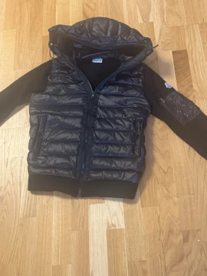 Cardigan - Snygg svart hybridjacka från Moncler med vadderad kropp och stickade ärmar. Jackan har huva, två framfickor med dragkedja och en extra ficka på ena ärmen med Moncler-logga. Perfekt för höst och vår! Använd men i fint skick.