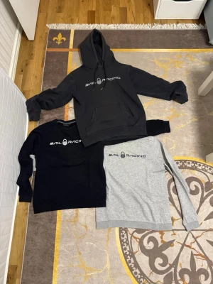 Tre Sail Racing hoodies i olika färger - Säljer tre snygga Sail Racing hoodies i svart, mörkgrå och ljusgrå. Den mörkgrå har huva och magficka, medan de andra två är klassiska sweatshirts utan huva. Alla har Sail Racing-loggan tryckt på bröstet. Perfekta för en avslappnad och sportig stil.