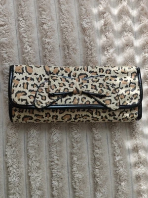 Kuvertväska med leopardmönster och rosett - Snygg kuvertväska i leopardmönstrat tyg med svart kant och en stor dekorativ rosett på framsidan. Väskan har en avlång form och stängs med lock. 