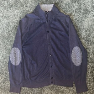 Marinblå cardigan från Tommy Hilfiger - Snygg marinblå cardigan från Tommy Hilfiger med knäppning framtill och ribbad krage i grått. Tröjan har mönstrade armbågslappar i grått och diskreta röda detaljer på insidan av kragen. Perfekt för en stilren look.