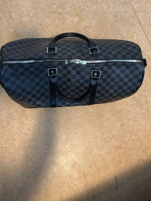 Svart weekendbag med rutmönster - Snygg svart weekendbag med klassiskt rutmönster och detaljer i svart läderimitation. Väskan har dubbla handtag, silverfärgade metalldetaljer och dragkedja upptill. Perfekt storlek för resor och weekendäventyr.