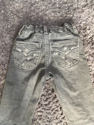 Grå jeans med broderade bakfickor - Säljer ett par grå jeans med raka ben och elastisk midja. Bakfickorna har snygga broderade detaljer och dekorativa knappar. Det är i storlek 158 från Gina, så det är för banr och tonåringar mest.