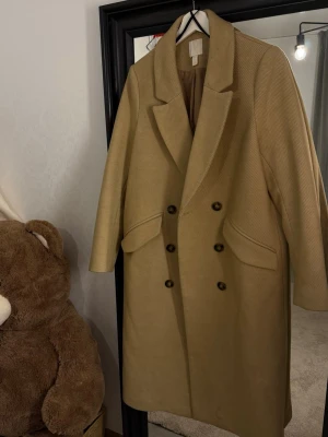Beige dubbelknäppt lång kappa HM - Säljer en stilren beige kappa med dubbelknäppning och breda slag. Kappan har två stora fickor framtill och är tillverkad i ett strukturerat tyg som ger en exklusiv känsla. Perfekt för dig som vill ha en klassisk och trendig look.